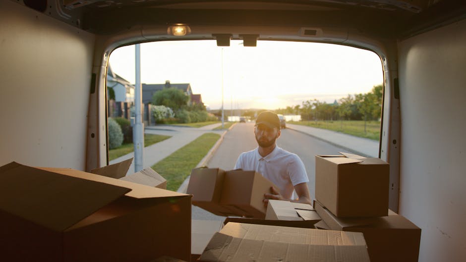 A man wearing a white polo shirt and a dark cap is standing outside a white moving van, holding a cardboard box in his hands during a home furniture transport process. The van's rear door is open, rev