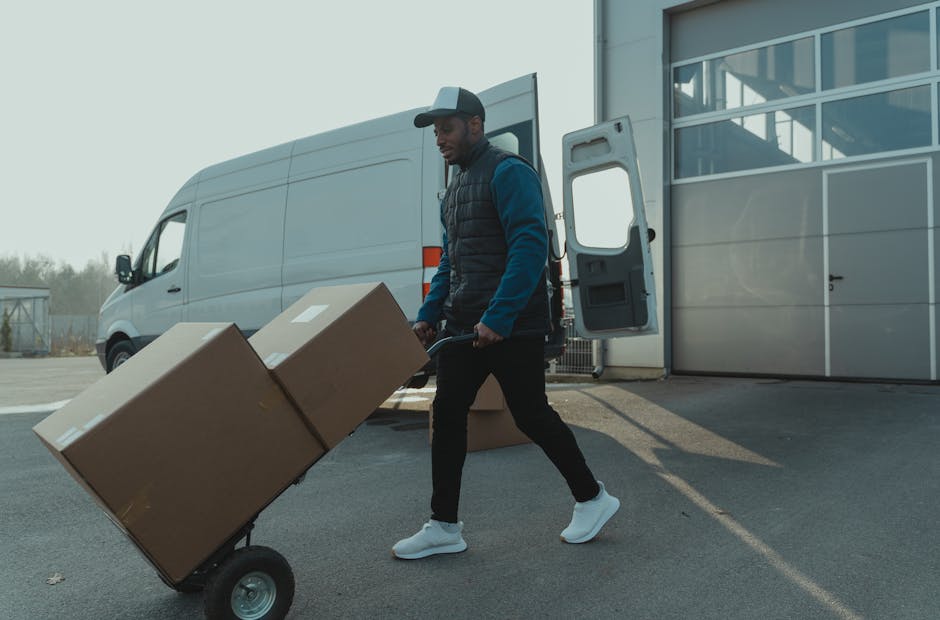 A man wearing a white polo shirt and a dark cap is standing outside a white moving van, holding a cardboard box in his hands during a home furniture transport process. The van's rear door is open, rev
