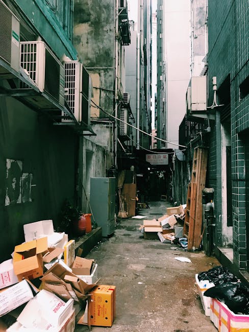 A narrow cobbled alleyway between two buildings, featuring brick paving and various external utility boxes, air conditioning units, and electrical wiring attached to the walls. On the right side, there are several large black wheelie bins lined up along the pavement, and an open doorway partially visible on the left. The environment appears to be part of an urban residential or commercial property, with limited natural light and a utilitarian atmosphere. This setting is representative of tight urban spaces often encountered during house removals or furniture transport in locations such as Hanwell, where navigating narrow streets and stairs is typical. Man with Van Hanwell’s removal services may utilize this area for loading or unloading furniture and boxes as part of a home relocation process involving careful stair and street access.
