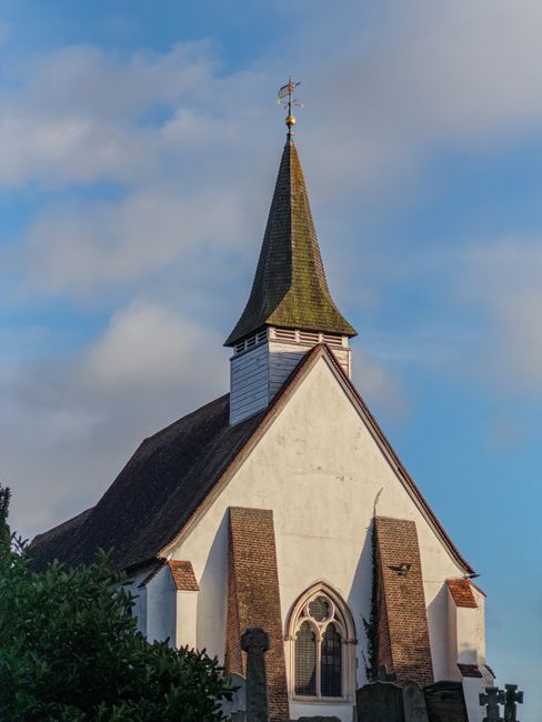 A white-painted church building with a steeply pitched roof and a tall, pointed spire topped with a weather vane, situated against a partly cloudy blue sky. The church features brick accents around arched windows and along the roofline, with some foliage visible at the bottom left corner. The architecture is traditional, and the spire extends prominently above the surrounding trees. The image reflects a clear and bright day, with natural lighting highlighting the structure's details. As part of house removals or relocation services, Man with Van Hanwell may use such an image to depict the types of buildings involved in local moving projects, emphasizing careful maneuvering around historic or traditional structures.