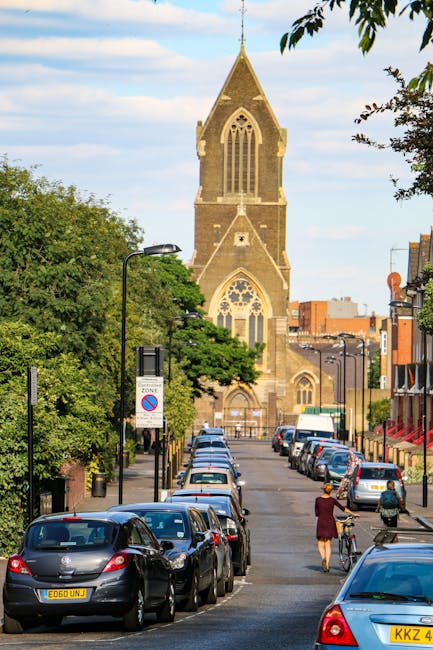 A white-painted church building with a steeply pitched roof and a tall, pointed spire topped with a weather vane, situated against a partly cloudy blue sky. The church features brick accents around arched windows and along the roofline, with some foliage visible at the bottom left corner. The architecture is traditional, and the spire extends prominently above the surrounding trees. The image reflects a clear and bright day, with natural lighting highlighting the structure's details. As part of house removals or relocation services, Man with Van Hanwell may use such an image to depict the types of buildings involved in local moving projects, emphasizing careful maneuvering around historic or traditional structures.