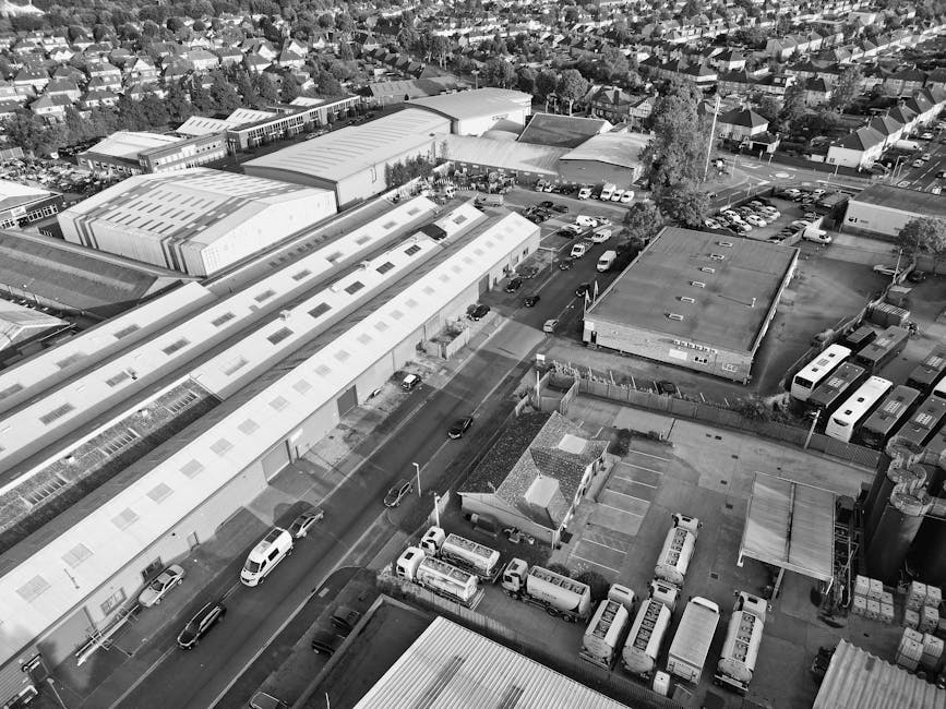 An aerial black-and-white photograph of a commercial and industrial area in Hanwell, showing multiple warehouse-style buildings with large flat roofs, surrounded by parking lots filled with cars and vans. In the foreground, a street runs through the area with several vehicles parked along the curb and some in motion. Adjacent to the buildings, there are smaller structures, possibly offices or storage units, alongside a narrow road and a few trees. The scene includes a loading zone where a van belonging to Man with Van Hanwell, a professional removals company, is positioned outside a warehouse, ready for a home relocation or furniture transport operation. The environment is well-lit, indicating daytime, and the overall setting reflects a busy logistics hub that supports packing, loading, and moving services for residential or commercial removals, aligned with local council permits for van loading bays in Hanwell.