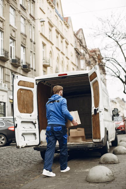 A man wearing a dark jacket and trousers is standing inside the open rear doors of a large white delivery van, which is parked on a paved street. The man is facing away from the camera, organizing or loading cardboard boxes of various sizes into the van's cargo space. The boxes are wrapped with labels and are stacked neatly, with some placed on a hand trolley positioned directly in front of the van, ready for transport or unloading. To the left of the van, a bicycle is partially visible, parked beside the paved area. The scene takes place during daytime, with trees and buildings visible in the background, suggesting an urban environment suitable for home relocation or furniture transport. The lighting appears natural, and the focus is on the process of packing or unpacking belongings as part of a professional moving service, such as that provided by Man with Van Hanwell, supporting efficient packing and loading in household removals.