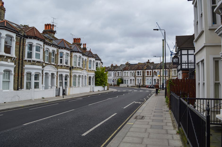 A quiet residential street scene showing a row of Victorian-style terraced houses with brick facades, white window frames, and pitched roofs with chimneys under a cloudy sky. The pavement on the right