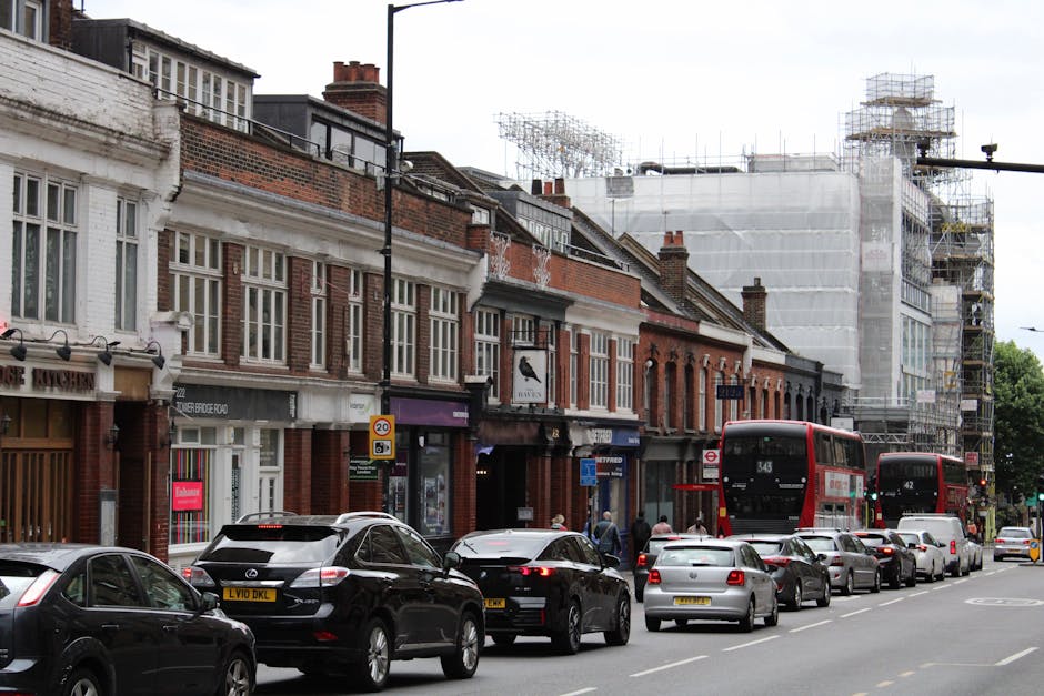 A street scene in Hanwell showing a row of brick and Victorian-style shops with large windows and signage, positioned along the pavement. There are multiple vehicles parked along the roadside, including a black Mercedes-Benz and a silver Kia, with additional cars visible behind them. Two red double-decker buses are also present, one numbered 343 and the other 42, heading in opposite directions. Pedestrians are walking along the pavement near the shops, some near the entrance of a store with a purple sign. In the background, a building under construction is covered with scaffolding and white protective sheeting, indicating ongoing development work. The scene is lit by daylight, with overcast skies, capturing a typical busy urban environment suited for house removals and furniture transport, as coordinated by Man with Van Hanwell.