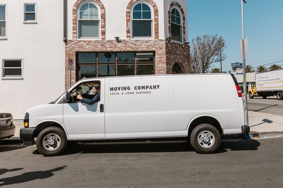 A man wearing a white t-shirt and jeans is standing on the pavement beside a white moving van with its rear door open. He is loading or unloading cardboard boxes, which are stacked on the ground in fr