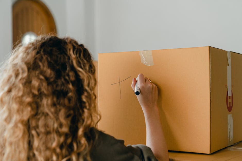 A woman with curly, light brown hair is seen from behind, writing the letter 'T' on a large cardboard moving box using a black marker. The box is positioned on a table or countertop inside a residential space, with visible packing tape securing it. The background shows a light-colored wall and a bronze or gold decorative mirror mounted on the wall. The scene depicts a typical home relocation process involving packing, with the woman marking the box for identification or labeling purposes. There are no other objects or furniture visible in the scene. This image is relevant to house removals and packing services provided by Man with Van Hanwell, illustrating the preparation stage of a furniture transport or home moving process.