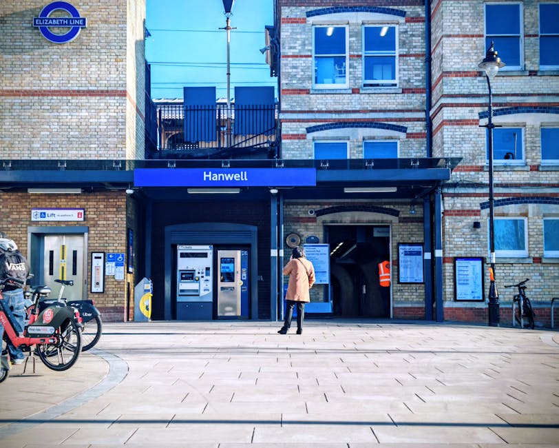 A woman standing outside the entrance of Hanwell station, which features a brick facade with large windows and a black canopy displaying the station name. To the left, there is a row of bicycles parked on a rack, including a dockless rental bicycle with branding visible. Next to the bicycles, several ticket machines and an automated kiosk are installed on the brick wall. The station entrance has a wheelchair-accessible lift control panel at ground level, with directional signage nearby, and a staircase leading up to the station platforms is visible behind a black gated railing. A street lamp and a black bollard are positioned on the right side of the station entrance, and the pavement in front is clean, paved with rectangular tiles. The scene represents an urban transit hub suitable for people involved in home relocation or furniture transport services, with clear cues for loading and moving logistics, often facilitated by moving specialists like Man with Van Hanwell.