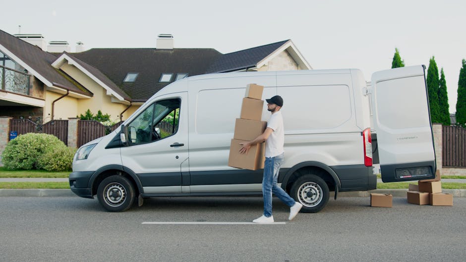 A man wearing a white T-shirt, blue jeans, white sneakers, and a black cap is walking alongside a white delivery van on a residential street in Hanwell, W7. He is carrying a stack of three cardboard boxes, gripping them with both hands as part of a home relocation or furniture transport process. The van, parked parallel to the curb, has its rear side door open, revealing a loading area for packing and move-related items. Several additional cardboard boxes are on the ground near the rear of the van, ready to be loaded or unloaded. The background features modern houses with pitched roofs, dormer windows, and well-maintained front gardens with bushes and trees, under bright daylight with clear weather. The scene captures an active loading process typical of professional removal services by Man with Van Hanwell, emphasizing the steps involved in packing, moving, and transporting household belongings.