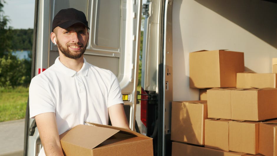 A man with a beard, wearing a black cap and a white polo shirt, is standing outside a moving van on a driveway or roadside, holding a cardboard box. The side door of the van is open, revealing several stacked cardboard boxes inside, some on the ground next to the vehicle. The boxes vary in size and are sealed with packing tape, suggesting a home relocation or furniture transport process. On the pavement near the van, there is a trolley or dolly that may be used for carrying heavier items, indicating a professional approach to packing and moving. The background includes greenery and clear skies, with natural lighting highlighting the scene and the man smiling at the camera. This image depicts the unloading or loading process typical of experienced removals services such as those offered by Man with Van Hanwell, emphasizing careful handling of household items during a house move.