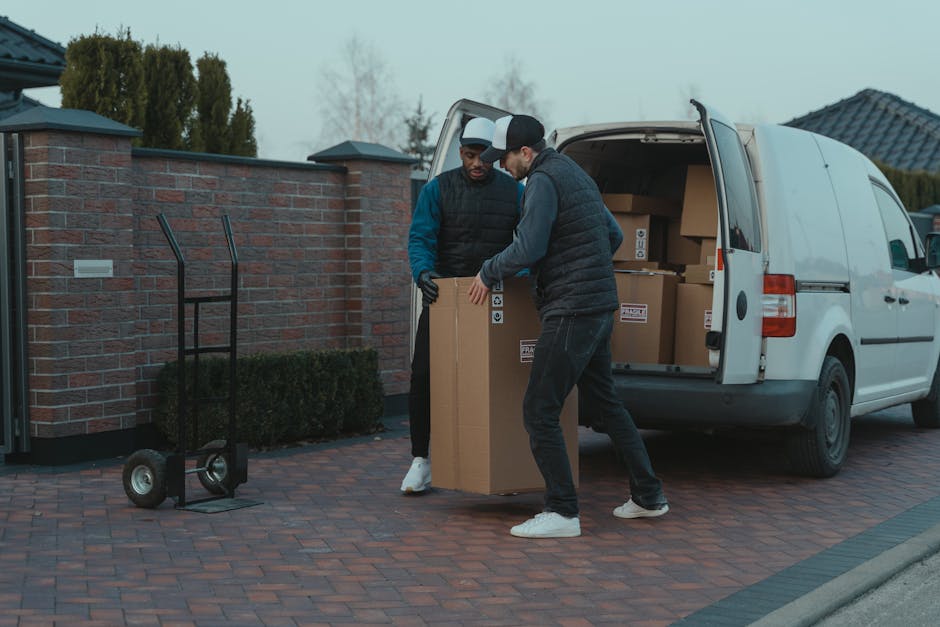 A man wearing a white T-shirt, blue jeans, white sneakers, and a black cap is walking alongside a white delivery van on a residential street in Hanwell, W7. He is carrying a stack of three cardboard boxes, gripping them with both hands as part of a home relocation or furniture transport process. The van, parked parallel to the curb, has its rear side door open, revealing a loading area for packing and move-related items. Several additional cardboard boxes are on the ground near the rear of the van, ready to be loaded or unloaded. The background features modern houses with pitched roofs, dormer windows, and well-maintained front gardens with bushes and trees, under bright daylight with clear weather. The scene captures an active loading process typical of professional removal services by Man with Van Hanwell, emphasizing the steps involved in packing, moving, and transporting household belongings.