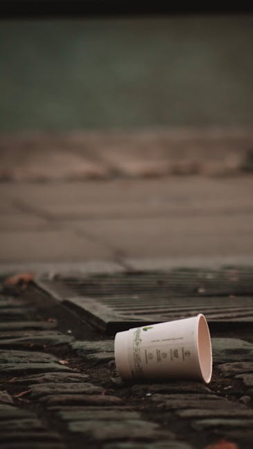 A close-up view of a hand holding a blue plastic rubbish bag filled with waste, with the bag's top gathered and twisted at the handle. The hand, partially visible, grips the bag firmly. The background